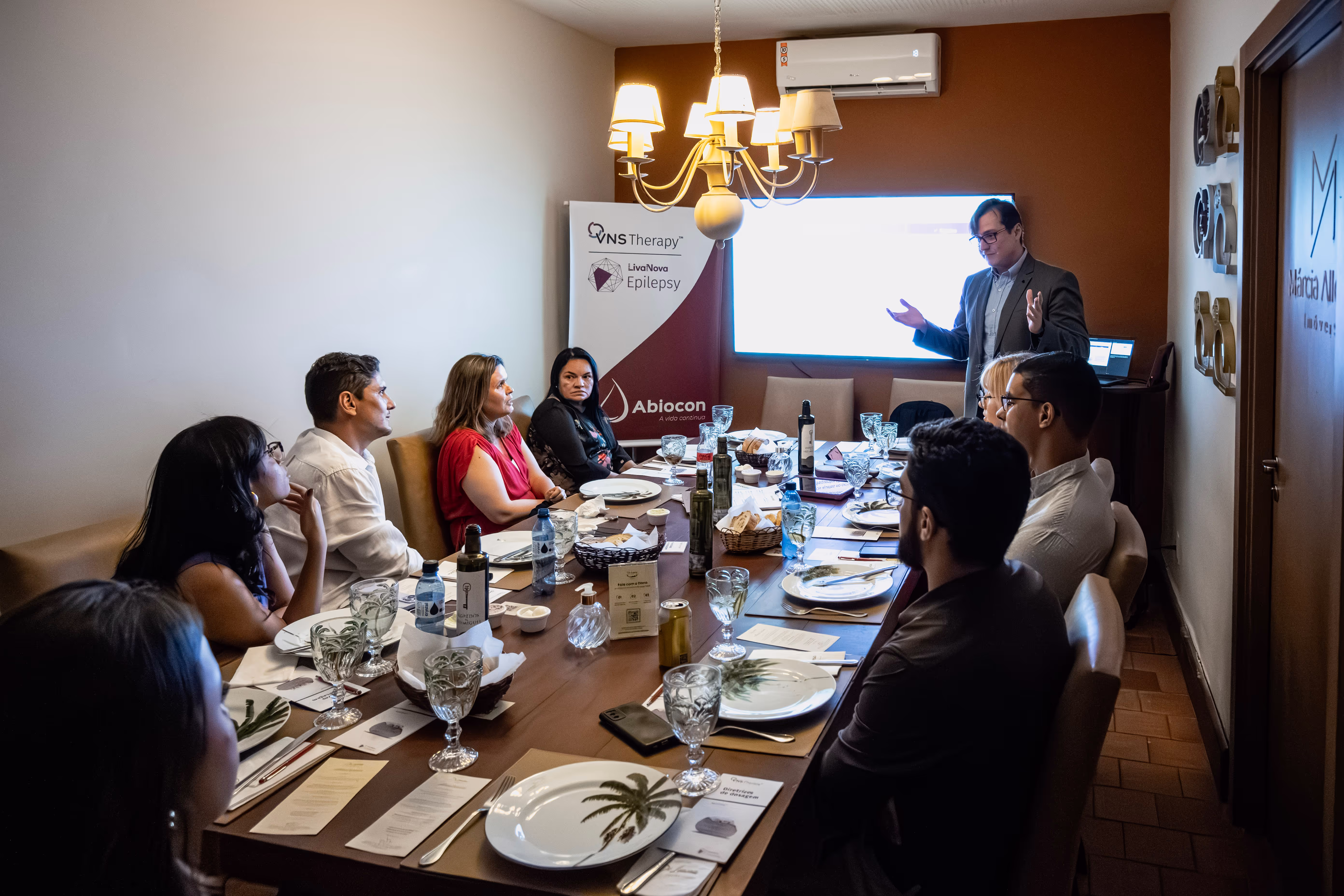 Encontro corporativo em um ambiente elegante, onde um grupo de profissionais está reunido em uma mesa de jantar enquanto assiste a uma apresentação. Um palestrante, de terno, gesticula enquanto explica o conteúdo exibido em uma tela iluminada ao fundo. O ambiente conta com decoração sofisticada e materiais de apresentação da Abiocon e da VNS Therapy LiveNow Epilepsy, sugerindo um evento sobre soluções médicas e inovação na área da saúde.