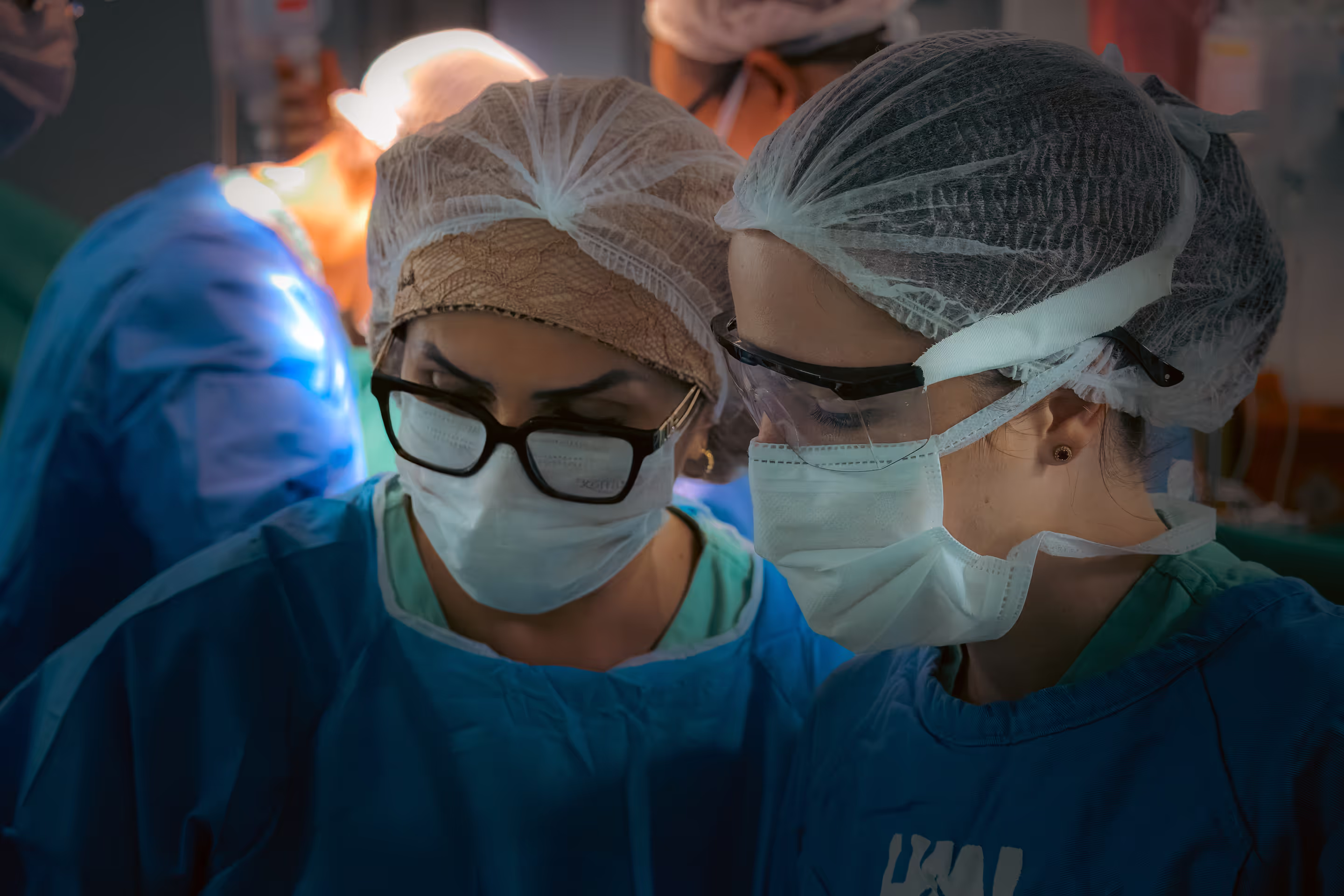 Duas profissionais da saúde em um ambiente cirúrgico. Ambas estão vestindo aventais cirúrgicos azuis, toucas descartáveis para cobrir os cabelos e máscaras faciais brancas. A pessoa à esquerda usa óculos de grau por cima da máscara, enquanto a da direita está usando óculos de proteção. Elas estão concentradas, olhando para baixo, possivelmente acompanhando ou realizando um procedimento médico. Ao fundo, desfocados, há outros profissionais também com vestimentas cirúrgicas, indicando que a cena ocorre dentro de uma sala de cirurgia. A iluminação é focada e reforça o ambiente técnico e sério do momento.