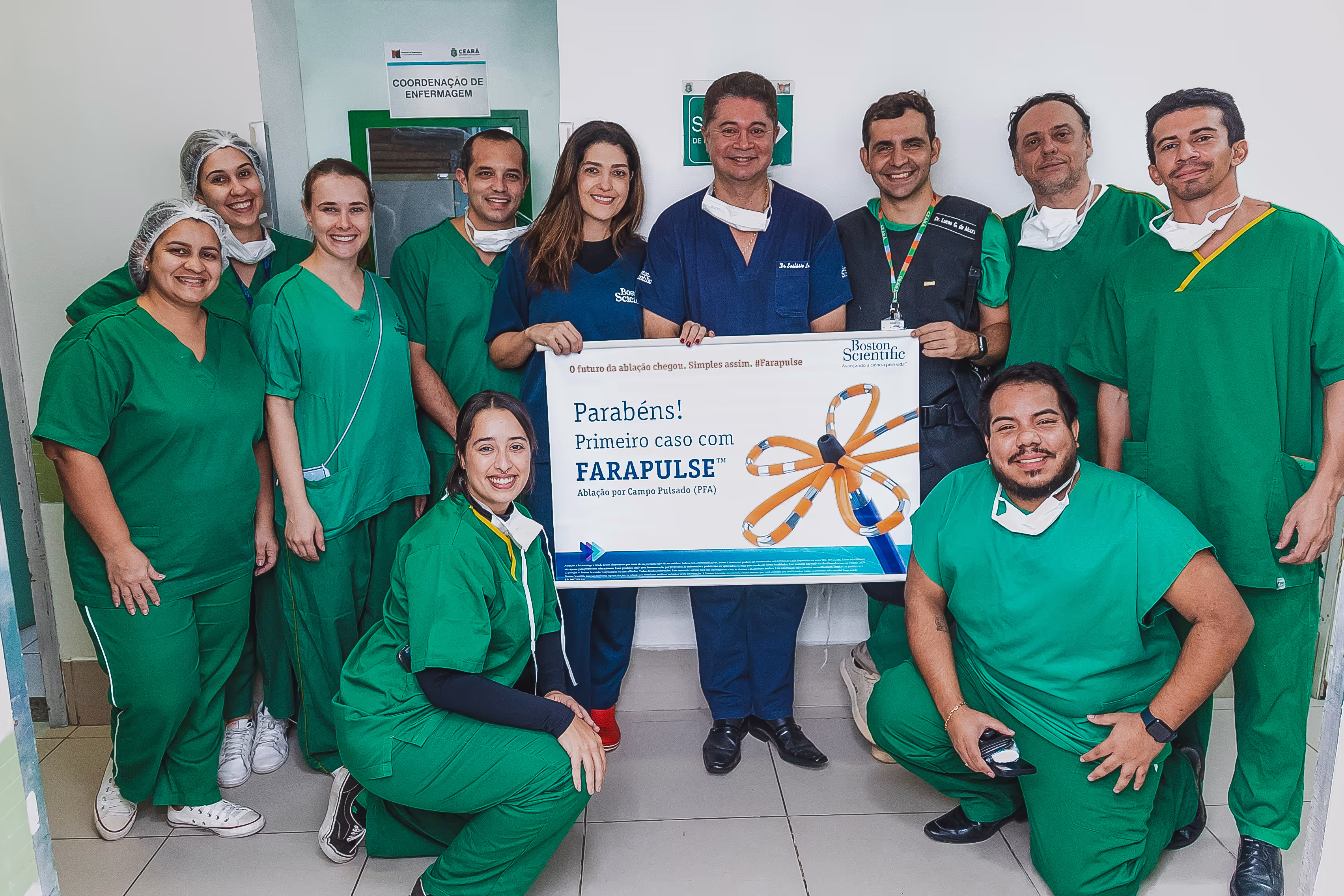 Equipe médica reunida para celebrar um marco na cardiologia. O grupo, composto por profissionais de saúde vestidos com jalecos e roupas cirúrgicas verdes, segura um cartaz anunciando o primeiro caso com FARAPULSE em Fortaleza-CE. O cartaz exibe a marca Boston Scientific e destaca a ablação por campo pulsado (PFA) como uma inovação no tratamento de arritmias. Todos na foto demonstram satisfação e orgulho pelo avanço tecnológico na área médica.