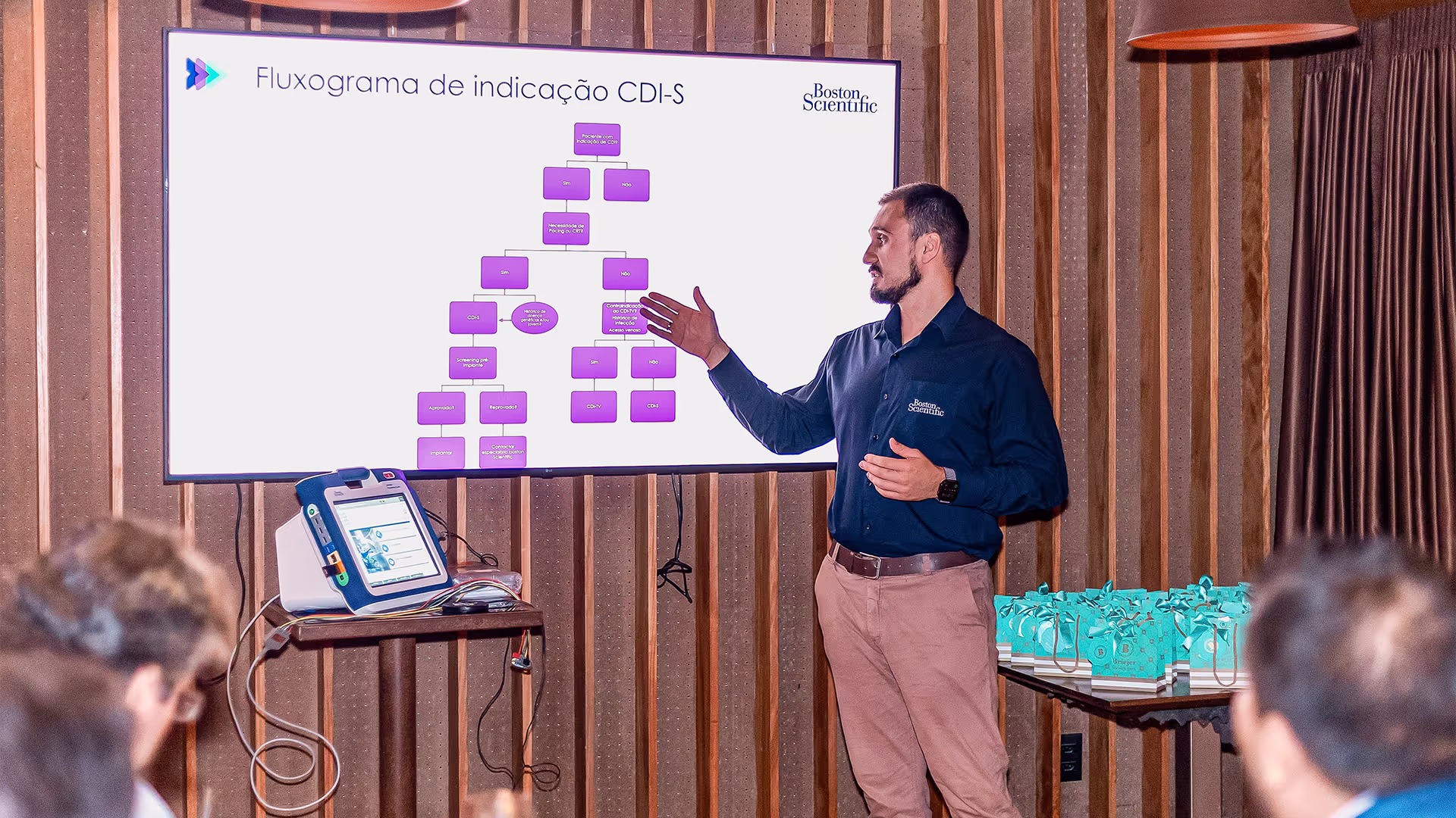 Um palestrante, vestindo uma camisa azul da Boston Scientific, apresenta um fluxograma de indicação do CDI-S em uma tela durante um evento. Ele gesticula em direção ao gráfico enquanto explica o conteúdo para a plateia. Ao lado da tela, há um dispositivo médico sobre uma mesa, conectado por cabos. No fundo, uma mesa exibe várias sacolas de brindes organizadas. O ambiente tem um design sofisticado, com iluminação aconchegante e paredes de madeira.